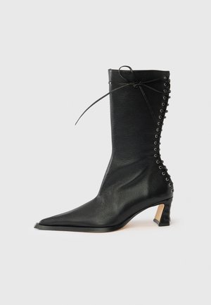 Ghete negre din piele, până la glezna, cu vârf ascuțit, toc subțire, detaliu cu șireturi în spate și ochiuri metalice. Textură netedă cu un design elegant.