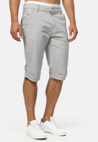 Homme portant un short écossais gris et blanc mi-long et des baskets blanches, debout devant un fond uni.
