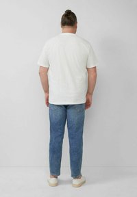 White short-sleeve t-shirt with a crew neck, worn with light blue denim jeans and white sneakers, viewed from the back.