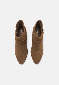 Botas de tobillo de ante marrón con punta afilada, diseño de correa envolvente y acento de hebilla dorada. Textura suave y silueta ajustada.