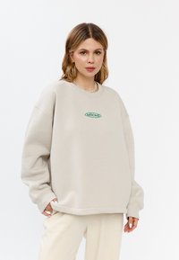 Lichtbeige sweatshirt met een oversized pasvorm, geribbelde manchetten en een afgeronde halslijn. Heeft een groene geborduurde logo in ovale vorm op de borst.