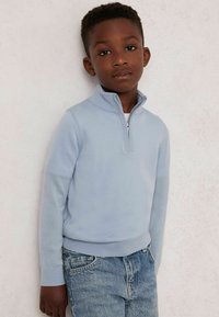 Gaiši zils kapuci džemperis, izgatavots no mīksta dzijas materiāla, ar garām piedurknēm un augstu apkakli. Pārī ar izbalējušām ziliem džinsiem.