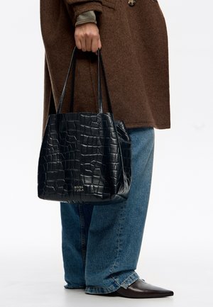 Personne tenant un sac fourre-tout en cuir noir texturé avec des doubles poignées, portant un manteau marron, un jean bleu et des chaussures pointues marron foncé.