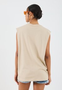 Beige ujjatlan póló, puha anyagból készült, kerek nyakkal és laza szabással, farmer sorttal és narancssárga napszemüveggel viselve.