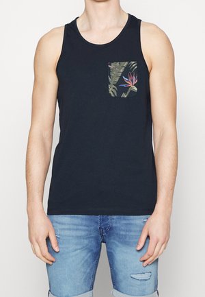 Mann trägt ein ärmelloses schwarzes Tanktop mit tropischem Blumenmuster auf der Brusttasche und hellblaue, hochgekrempelte Jeansshorts.