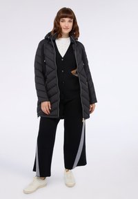 Chaqueta de plumas negra con capucha, diseño acolchado, cierre frontal, llevada sobre un suéter negro y pantalones de pierna ancha con rayas gris en los laterales. Zapatillas blancas.