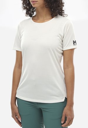 Frau trägt ein weißes, kurzärmeliges Sportshirt und grüne Hose, steht vor einem schlichten weißen Hintergrund.