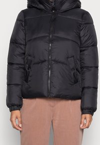 Czarna kurtka puchowa z kapturem, poziomy wzór pikowania, pełny zamek, elastyczne mankiety i dwie boczne kieszenie. Nosi się ją na beżowych spodniach.