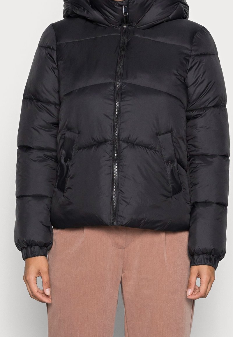 Czarna kurtka puchowa z kapturem, poziomy wzór pikowania, pełny zamek, elastyczne mankiety i dwie boczne kieszenie. Nosi się ją na beżowych spodniach.