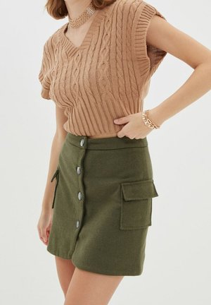 Femme portant un pull sans manches en maille torsadée beige et une jupe boutonnée kaki avec poches plaquées, accessoirisée avec des bijoux en or.