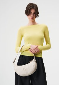 Beige halbmondförmige Tasche mit einem Reißverschluss, verziert mit zwei farbigen Punkten als Akzent. Modell trägt ein langärmeliges gelbes Oberteil und schwarze Hose.