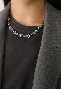 Collar de cadena de plata con eslabones redondos y ovalados, llevado sobre una camiseta gris oscuro debajo de un blazer gris con rayas finas.