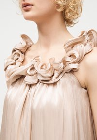 Γυναίκα με κοντά, σγουρά ξανθά μαλλιά, φορώντας ένα ανοιχτό μπεζ πλισέ φόρεμα με μεγάλα, βολάν από σατέν λουριά γύρω από το ντεκολτέ.