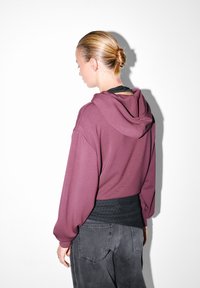 Žena s blond vlasmi v drdole, oblečená v fialovej mikine, čiernom perforovanom tričku a tmavošedých džínsoch, stojí proti bielej stene v bočnom profile.