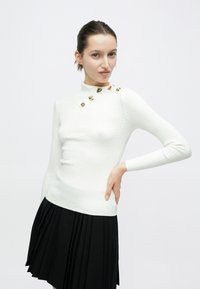 Λευκό πλεκτό πουλόβερ με ριμπ ύφανση και ψηλό γιακά, με χρυσές λεπτομέρειες σε έναν ώμο. Συνδυάζεται με μαύρη πλισέ φούστα.