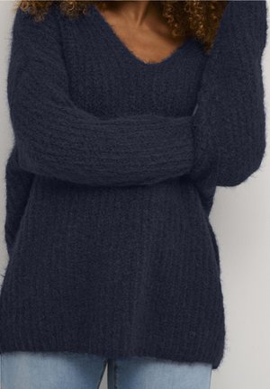 Personne portant un pull en maille duveteux bleu marine à manches longues et un jean bleu, les bras croisés sur la poitrine.