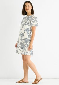 Short-sleeve dress in cream fabric with navy floral print. Features puffed sleeves and falls above the knee. Worn with brown flat sandals.