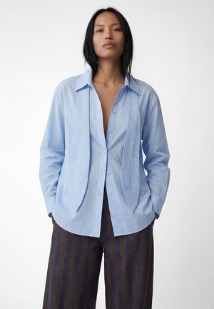 Kvinde med langt sort hår iført en lyseblå skjorte med smalle striber og mørke, vidbenede bukser med striber mod hvid baggrund.