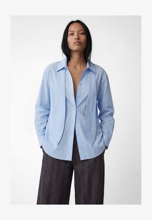 Femme aux longs cheveux noirs portant une chemise boutonnée bleu clair à fines rayures et un pantalon large à rayures foncées, sur fond blanc.