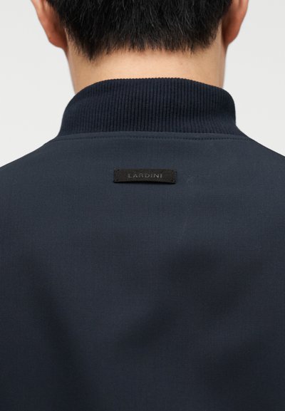 Μπλε ναυτικό μπουφάν με ριμπ γιακά, που φέρει μια μαύρη υφασμάτινη ετικέτα με την επιγραφή "LARDINI" στο πίσω μέρος, με λεία υφή και ραμμένη σχεδίαση.