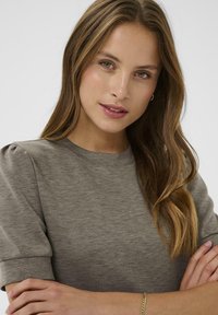 Siva majica kratkih rukava s okruglim izrezom i suptilnim napuhanim rukavima, izrađena od meke teksturirane tkanine, nosi se uz jednostavnu narukvicu.