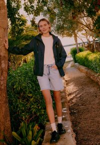 Jeune femme en veste noire, t-shirt blanc et short en jean, debout, une main posée sur un arbre et l'autre dans sa poche, sur un chemin de jardin.
