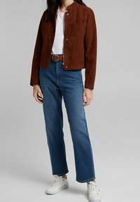 Bruine suède jas met een mandarin kraag, twee voorknosjes, gecombineerd met blauwe rechte jeans en witte sneakers.