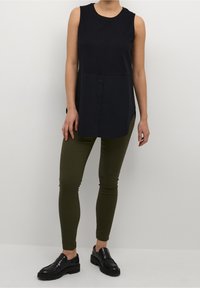 Schwarzes ärmelloses Oberteil mit Knopfleiste, kombiniert mit olivgrünen Leggings und schwarzen Slippern, die eine strukturierte Oberfläche und eine schlanke Passform aufweisen.