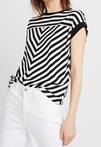Černobíle pruhované tričko s vzorem chevron, s krátkými ohrnutými rukávy, kulatým výstřihem a volným střihem, zkombinované s bílými džíny.