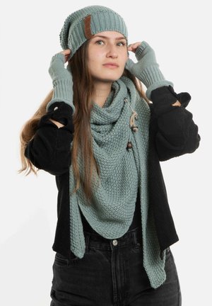 Bonnet en tricot et écharpe en teal doux, accompagnés de gants sans doigts. L'écharpe présente un motif texturé et un bouton décoratif. Le modèle porte les accessoires.