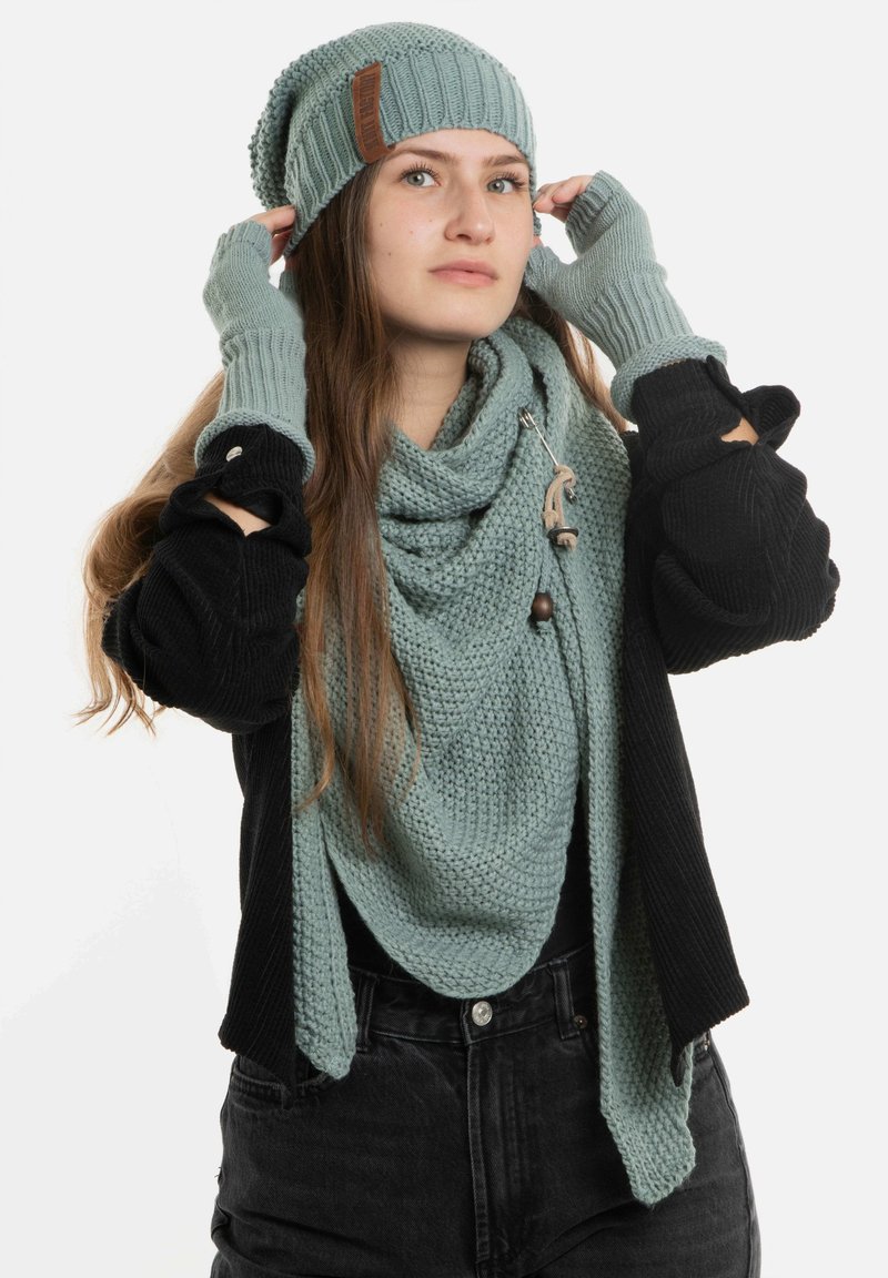 Gebreide beanie en sjaal in zacht teal, gecombineerd met vingerloze handschoenen. De sjaal heeft een textuurpatroon en een decoratieve sluiting. Model toont de accessoires.