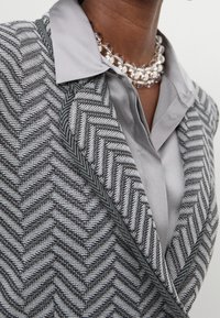 Blazer en motif chevron gris avec un col aiguisé, associé à une chemise grise en soie et un collier de perles superposé.