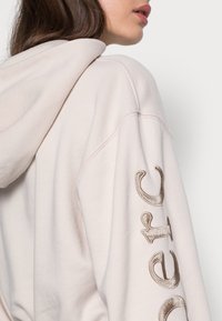 Světle růžová mikina s kapucí a vyšitým textem v metalické zlaté barvě na rukávu. Měkká, jemně kartáčovaná látka; volný střih s nastavitelnou kapucí.