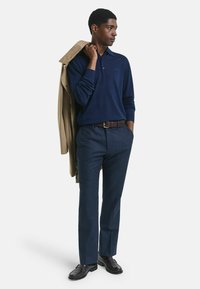 Man in navy lange mouw polo en blauwe broek met een beige jas over schouder, met één hand in de zak, staande tegen een witte achtergrond.