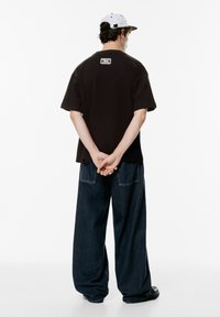 Sort kortærmet t-shirt, oversized pasform, med et logo på den øverste del af ryggen. Sammen med løse, mørke denimjeans og en grå kasket.