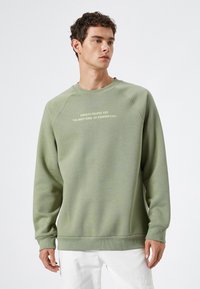 Sudadera verde con un cuello redondeado y mangas raglán largas. Presenta texto en un color más claro en el pecho. Textura suave.