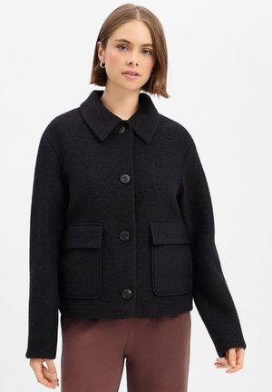 Woman wearing a black buttoned wool jacket with large front pockets and brown pants against a plain white background.