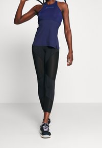 Sportovně založená osoba v námořnickém tílku a černých legínách s síťovými panely, která drží tenisovou raketu za hlavou.