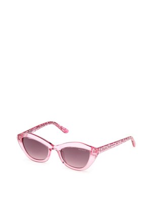 Des lunettes de soleil roses en forme d'œil de chat, avec des montures transparentes, des verres dégradés gris et des branches à motifs portant le nom de la marque "GUESS."