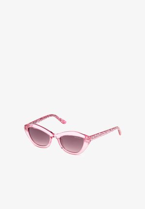 Des lunettes de soleil roses en forme d'œil de chat, avec des montures transparentes, des verres dégradés gris et des branches à motifs portant le nom de la marque "GUESS."