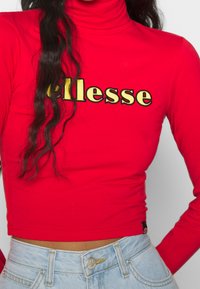 Rotes Langarm-Turtleneck-Oberteil mit "ellesse"-Logo in goldenen Buchstaben. Figurbetontes Design, weicher Stoff, kombiniert mit hellblauen, hochtaillierten Jeans.