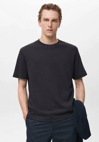 Tricou cu mânecă scurtă de culoare gri închis, cu decolteu rotund, având un material texturat și o croială lejeră. Purtați cu pantaloni închiși la culoare.