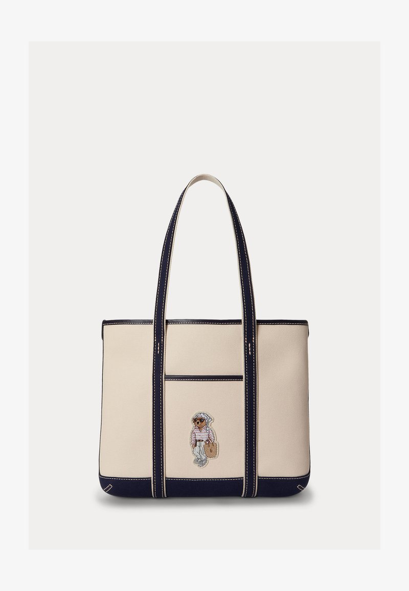 Beige and navy tote bag with long handles, navy trim, and an embroidered bear wearing sunglasses and a sailor outfit on the front pocket.