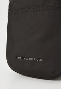 Crna tkanina torba preko ramena s glatkom teksturom, koja sadrži prednji džep i srebrno otisnuti logo "TOMMY HILFIGER" na dnu.