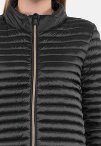 Giubbotto a vento nero con texture trapuntata, colletto alto e zip frontale con accenti dorati. Il materiale appare lucido e leggero.