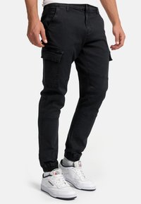 Bărbat purtând pantaloni jogger cargo negri și adidași albi Reebok, stând pe un fundal simplu, luminos.