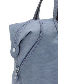 Světle modrý látkový batoh s výraznou zipovou kapsou a kulatým detailem knoflíku. Má texturovaný materiál a robustní černé popruhy.