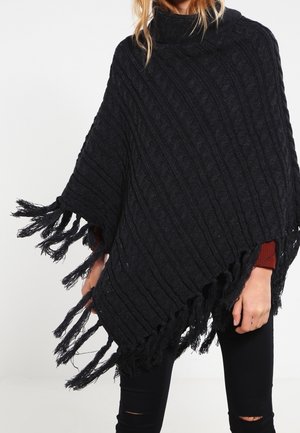 Donna che indossa un poncho nero lavorato a maglia con bordi sfrangiati sopra una maglia rossa a maniche lunghe e jeans neri strappati, in piedi davanti a uno sfondo bianco.