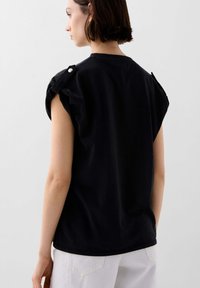 Black cotton t-shirt with short sleeves, featuring decorative shoulder buttons, smooth texture, and a relaxed fit. Background is light gray.