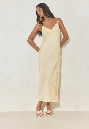 Kvinde iført en cremefarvet slipkjole med blondekant og tynde stropper og blomstret øreringe, poserer med hænderne bag ryggen.